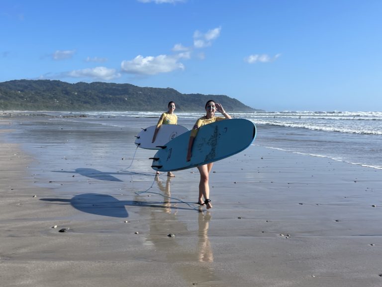 Two women in yellow rash guards walking along Santa Teresa beach carrying blue surfboards